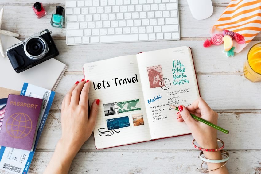 Person writing a travel planning checklist in a journal with passport, boarding passes, and camera on a desk.